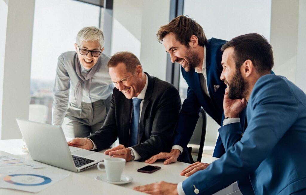 Group of business professionals smiling and collaborating around a laptop during a meeting in a modern office setting.